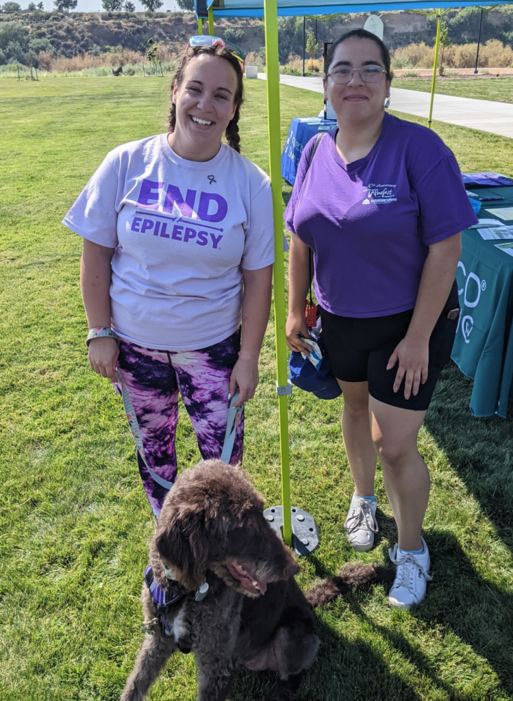Two women smiling at an outdoor epilepsy awareness event, one wearing an “END Epilepsy” shirt, with a large brown dog sitting in front of them.
