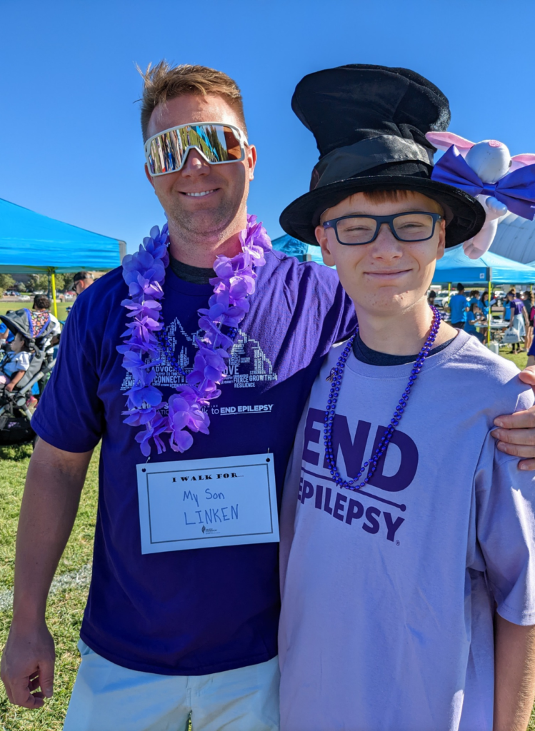 Two people smiling together outdoors at an epilepsy walk, wearing purple shirts and accessories under a bright blue sky.
