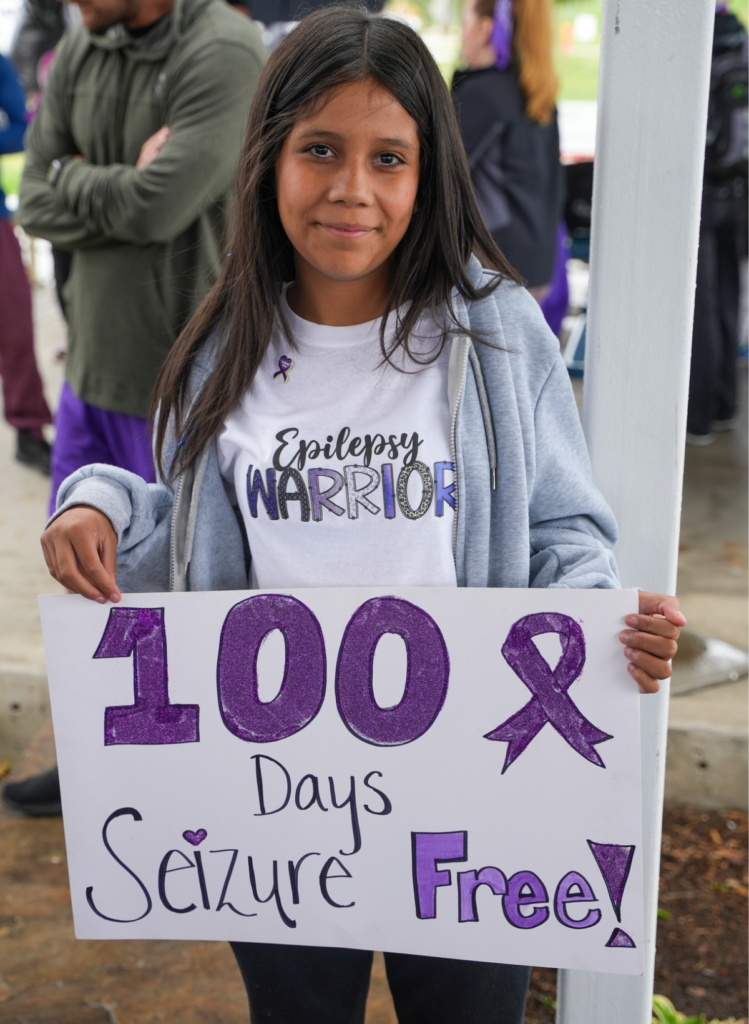 Young person wearing an “Epilepsy Warrior” shirt holds a handmade sign celebrating 100 days seizure-free, decorated with a purple epilepsy awareness ribbon.
