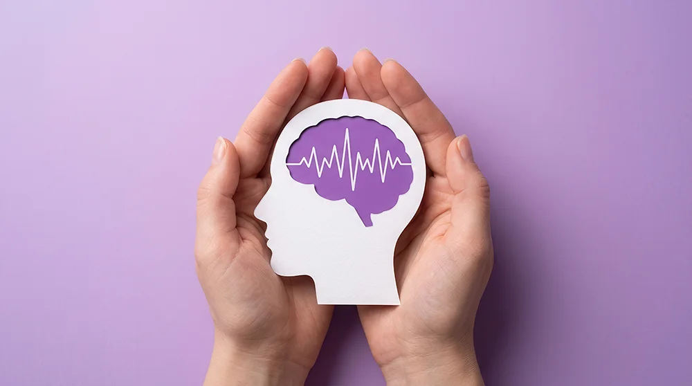 Hands gently cradle a paper silhouette of a human head with a purple brain and heartbeat line, symbolizing mental health care and support.
